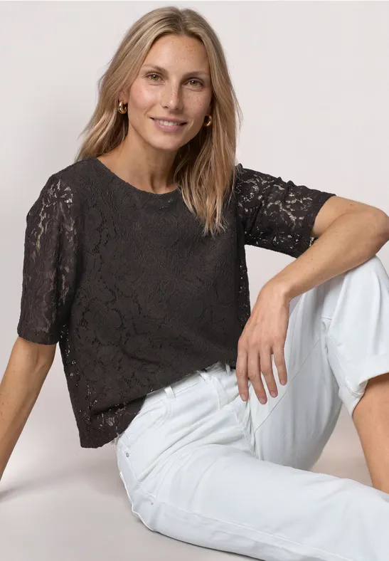 Frau sitzt und trägt schwarze Spitzenbluse mit kurzen Ärmeln und weiße Jeans.