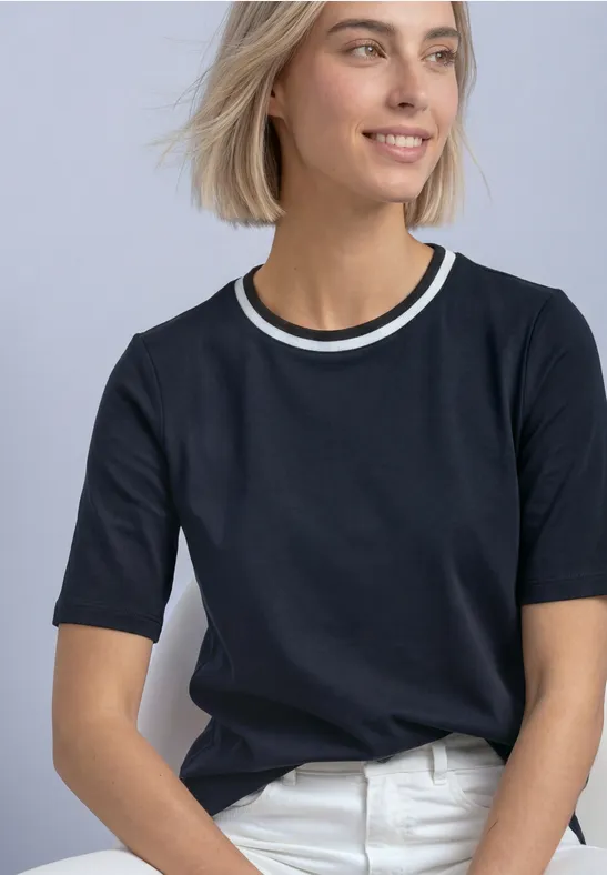 Młoda kobieta siedzi, uśmiechnięta, spogląda bokiem, ma czarny t-shirt z podwójnym białym kołnierzem i białe jeansy.