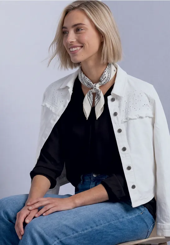 Eine blonde Frau sitzt lächelnd, trägt weiße Jeansjacke über schwarzem Oberteil, Jeanshose und ein Halstuch.