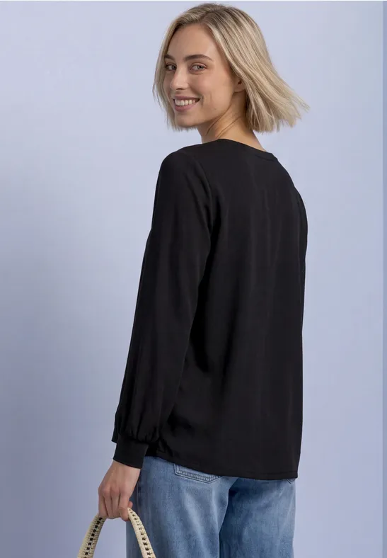 Frau mit kurzen blonden Haaren, trägt schwarzen Pullover und blaue Jeans, steht seitlich und schaut über die Schulter.