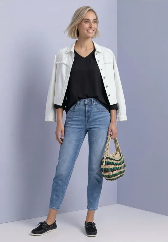 Junge Frau mit weißer Jacke, schwarzem Top, blauen Jeans, schwarzen Loafers und gestreifter Strandtasche.