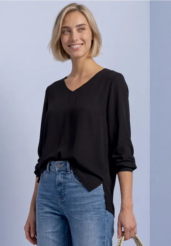Frau mit schwarzer Bluse und blauen Jeans, lächelnd vor hellgrauem Hintergrund.