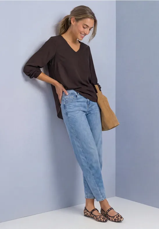 Junge Frau lehnt an heller Wand, trägt schwarzen V-Ausschnittpullover, helle Jeans, Leo-Sandalen und Schultertasche.