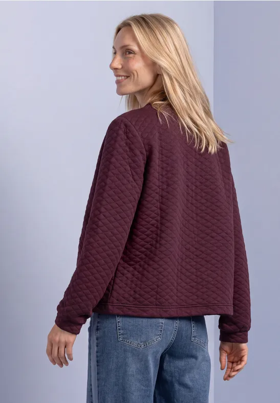 Eine Frau mit blonden Haaren trägt eine kastanienbraune Steppjacke und Jeans vor hellem Hintergrund.