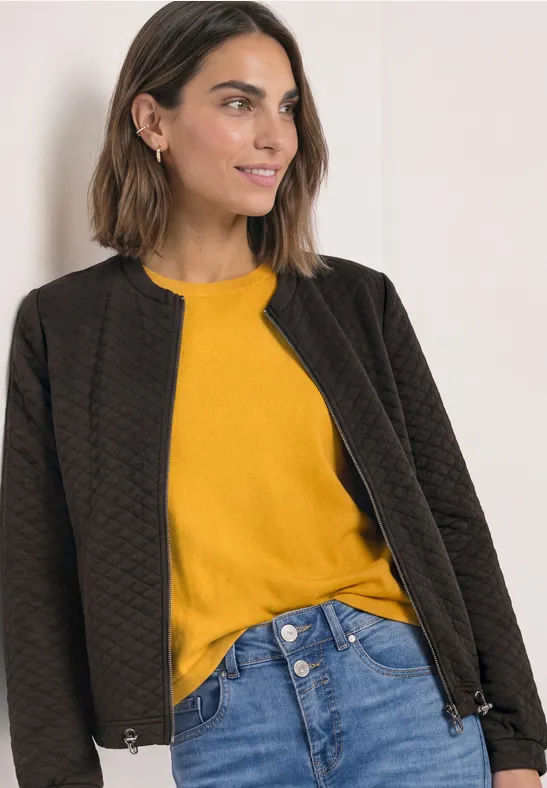 Eine Frau trägt eine gesteppt schwarze Jacke, ein gelbes Oberteil und blaue Jeans, leicht lächelnd.