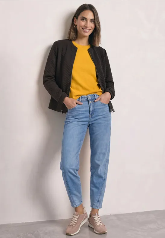 Frau trägt gelbes Oberteil, schwarze Jacke, blaue Jeans, beige Sneaker. Steht lächelnd vor neutraler Wand.
