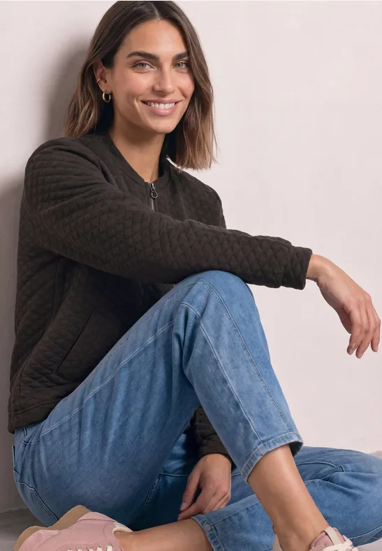 Junge Frau sitzt lässig auf dem Boden, trägt Jeans und dunklen Pullover und lächelt entspannt.
