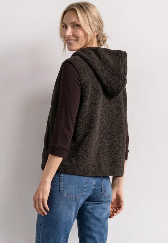 Eine Frau trägt eine schwarze Kapuzenweste über einem dunklen Langarmshirt und Jeans, schaut über die Schulter.