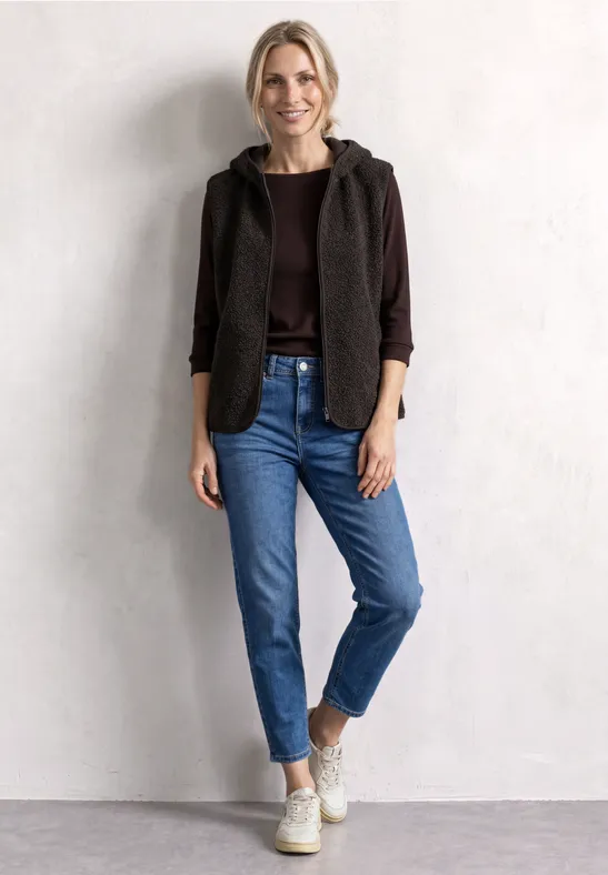 Junge Frau steht vor heller Wand, trägt braune Jacke mit Kapuze, braunes Shirt, blaue Jeans und weiße Sneakers.