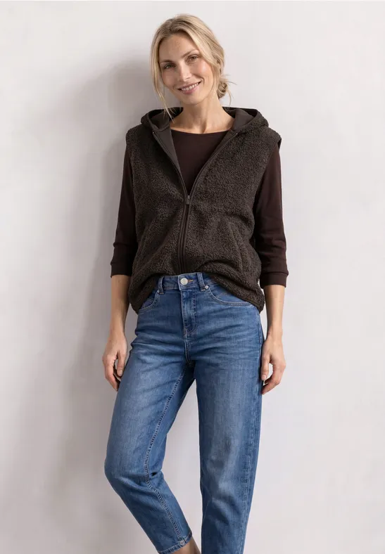 Eine Frau trägt eine braune Kapuzenweste über dunkelgrauem Shirt und blauen Jeans vor heller Wand.