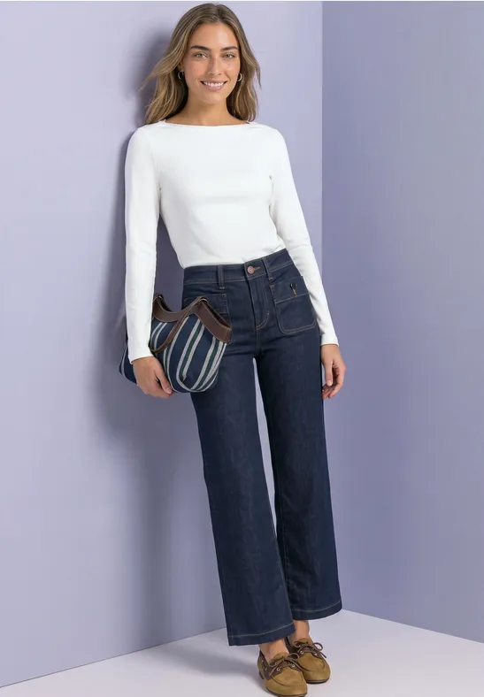Frau in weißem Langarmshirt, dunklen Jeans und Tasche vor violettem Hintergrund.