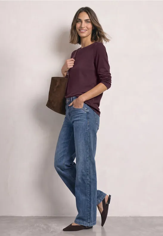 Vrouw in casual look met wijde jeans, bordeaux trui en een bruine tas.