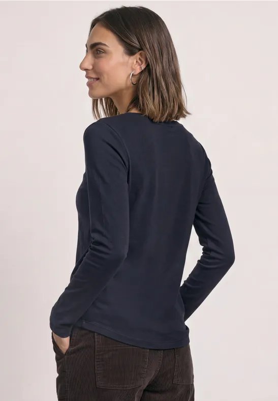 Kobieta widziana z profilu ma na sobie granatowy longsleeve z długim rękawem i brązowe spodnie, ręce w kieszeniach.