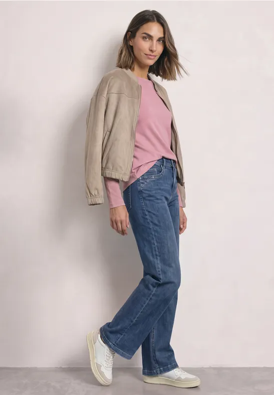 Eine Frau lehnt an einer Wand, trägt einen beigen Blazer über pinkem T-Shirt, weite Jeans und weiße Sneaker.