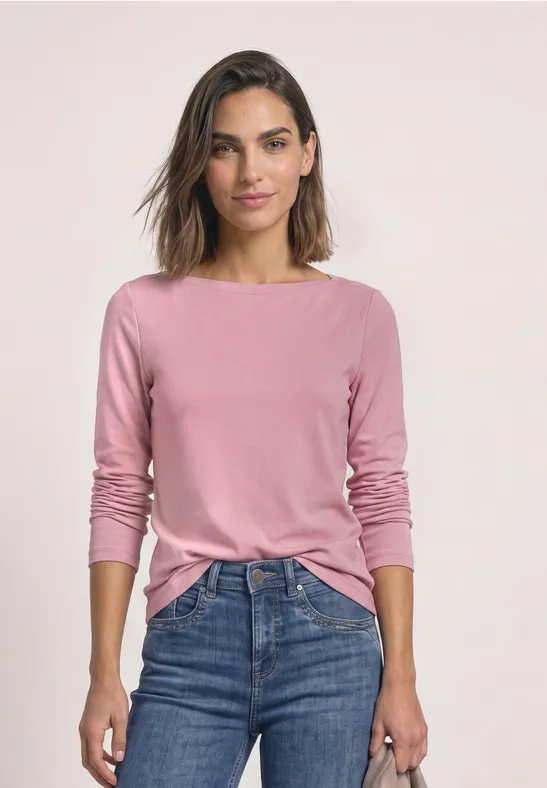 Frau mit rosafarbenem Langarmshirt und blauen Jeans steht frontal vor neutralem Hintergrund.