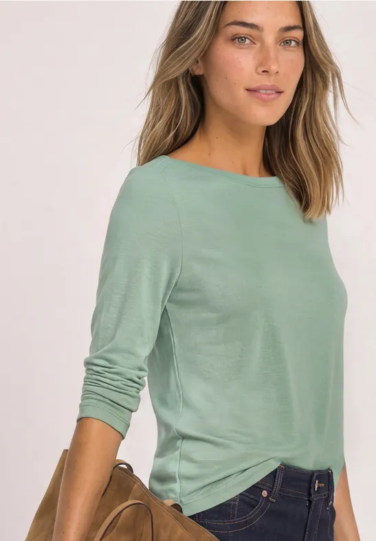Frau in hellgrünem Langarmshirt und Jeans, lässig vor hellem Hintergrund.