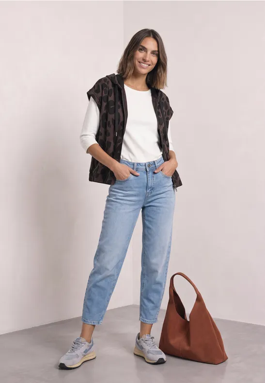 Frau in weißem T-Shirt, hellblauen Jeans, lässigen Sneakern und brauner Tasche vor hellem Hintergrund.