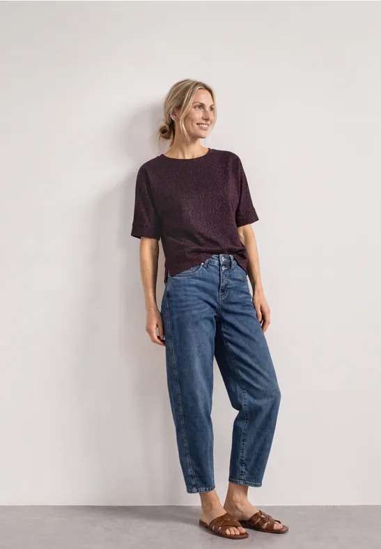 Eine lächelnde Frau lehnt an einer hellen Wand, trägt lila Shirt, hochtaillierte Jeans mit Umschlag und braune Sandalen.
