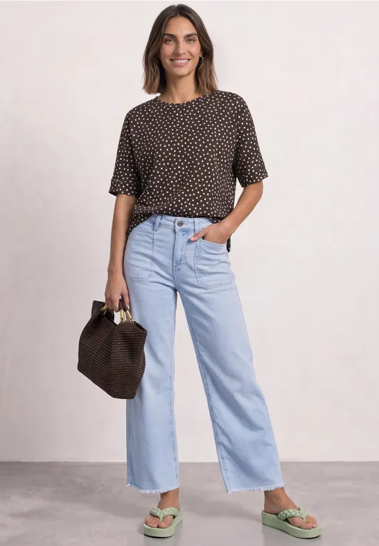 Frau in gepunktetem T-Shirt, hellblauen, weiter geschnittenen Jeans und grünen Sandalen mit brauner Tasche.