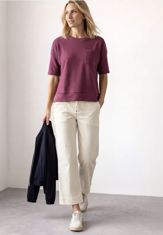 Une femme porte un t-shirt violet, un pantalon ample clair et des baskets blanches, elle tient une veste foncée.