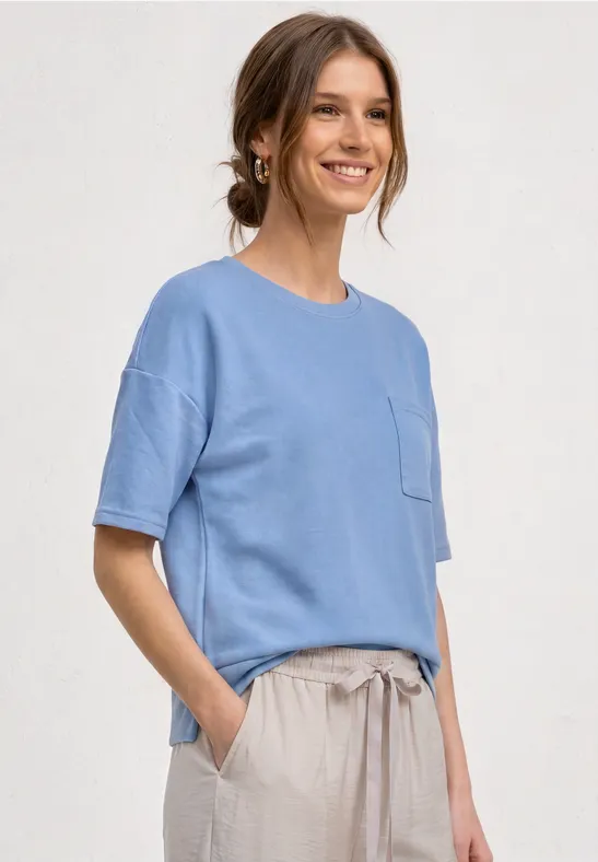 Een glimlachende vrouw in een blauw T-shirt en beige broek, handen in haar zakken.