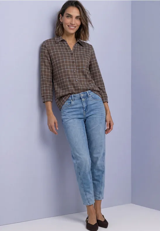 Frau in braun-karierter Bluse und hellblauen Jeans vor lila Hintergrund.