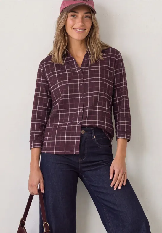 Eine junge Frau lächelt, trägt eine karierte Bordeaux-Bluse, dunkle Jeans, Schirmmütze und hält eine Tasche.
