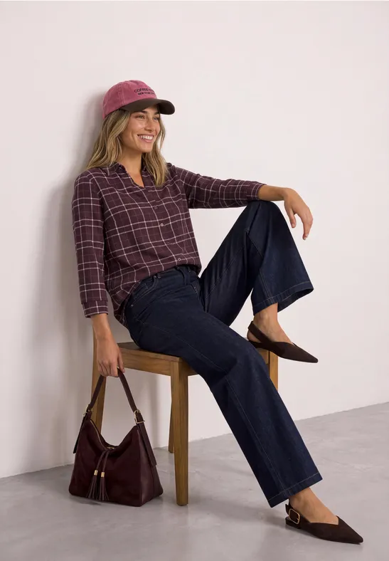 Frau mit rosa Cap, kariertem Hemd, weiter Jeans und Tasche sitzt entspannt auf Stuhl.