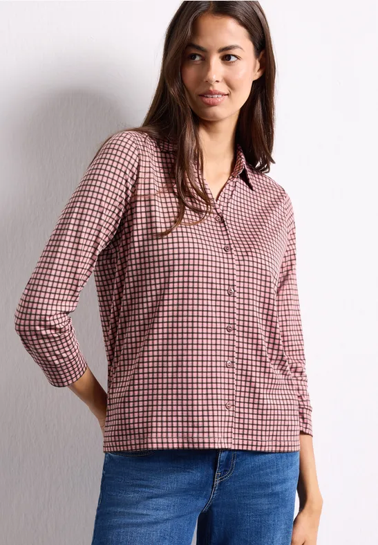 Eine Frau trägt eine rot-weiß karierte Bluse und blaue Jeans vor einer weißen Wand.