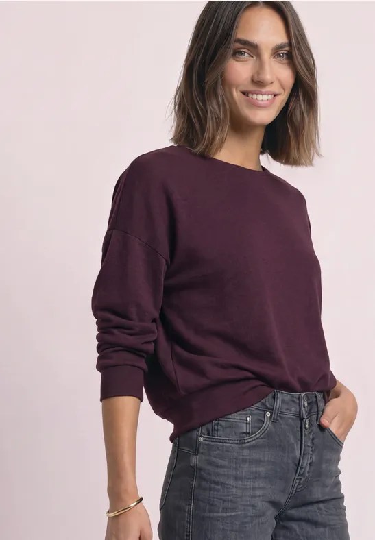Eine lächelnde Frau in einem dunkelroten Pullover und grau-blauen Jeans vor hellem Hintergrund.