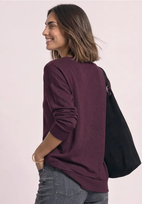 Frau in seitlicher Ansicht mit weinrotem Pullover, Jeans und schwarzer Tasche, lächelnd.