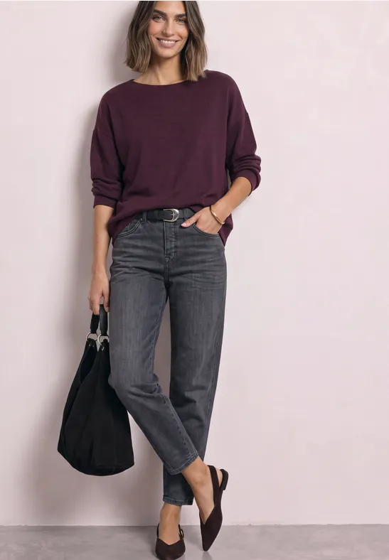 Frau trägt weinroten Pullover, graue Jeans, braune Loafers und hält schwarze Tasche vor hellgrauer Wand.