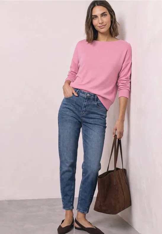 Vrouw in roze trui, blauwe jeans en zwarte ballerina’s met een bruine tas.