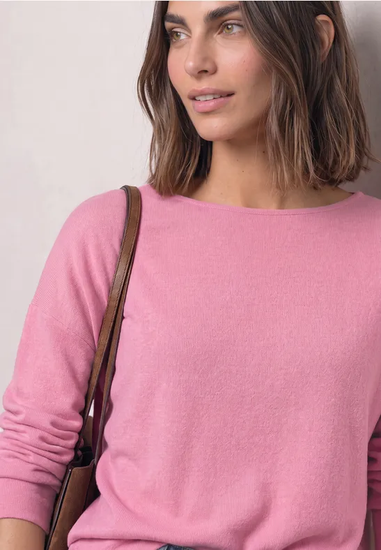 Vrouw met bruin haar in een roze trui en schoudertas, glimlachend, kijkt zijwaarts.