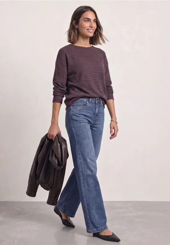 Eine Frau trägt eine gestreifte langärmlige Bluse, blaue Jeans und hält eine Jacke in der Hand.