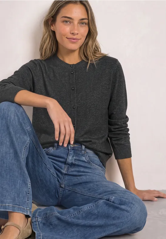 Eine lächelnde Frau in dunkelgrauem Cardigan und blauen Jeans sitzt entspannt vor hellem Hintergrund.