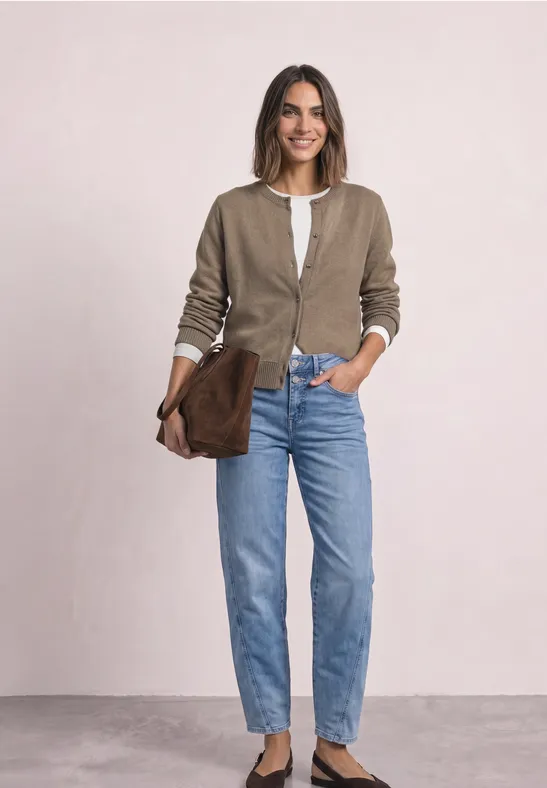 Frau lächelt, trägt beige Cardigan, weißes Top, blaue Jeans und hält eine braune Clutch vor hellem Hintergrund.