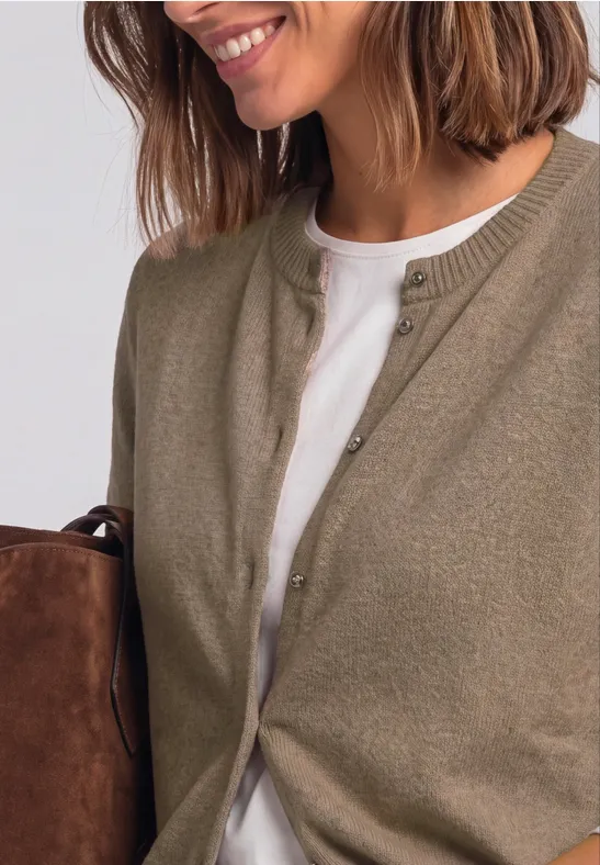 Frau mit schulterlangem braunem Haar trägt einen khakifarbenen Cardigan über weißem T-Shirt und hält eine Tasche.