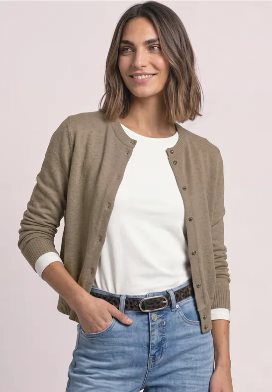 Eine lächelnde Frau in Jeans, weißem Shirt und brauner Strickjacke vor hellem Hintergrund.
