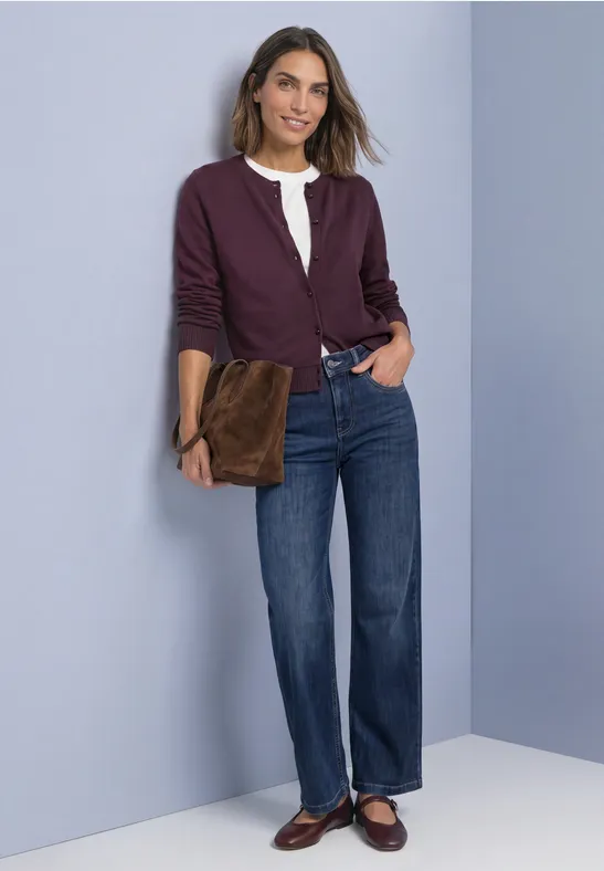 Femme souriante en blouse blanche, gilet violet, jean droit et mocassins marron, tenant une pochette marron.