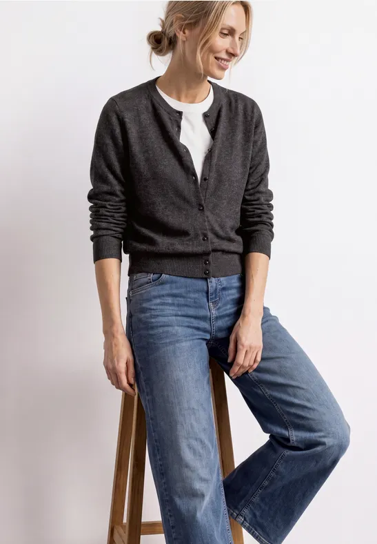 Frau lehnt lächelnd an einem Hocker, trägt grauen Strickcardigan, weißes Shirt und weite blaue Jeans.