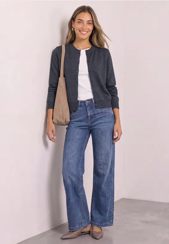 Frau lächelt in Jeans, weißem Shirt und dunkelgrauer Jacke mit Schultertasche gegen weiße Wand.