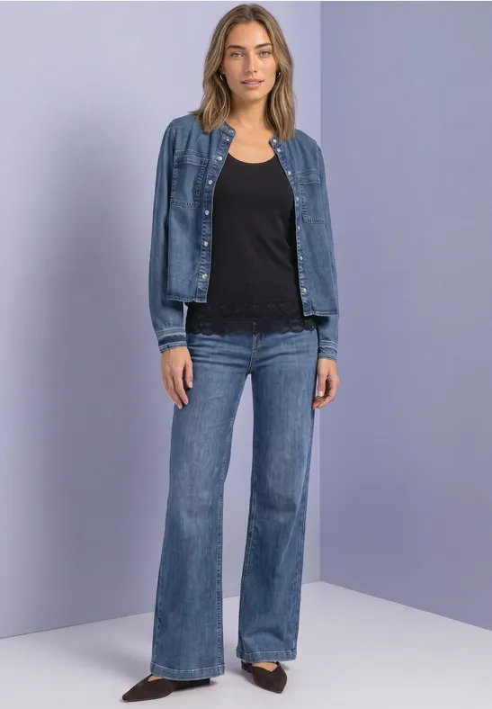 Eine Frau in schwarzer Bluse, Jeansjacke und ausgestellten Jeans vor neutralem Hintergrund.