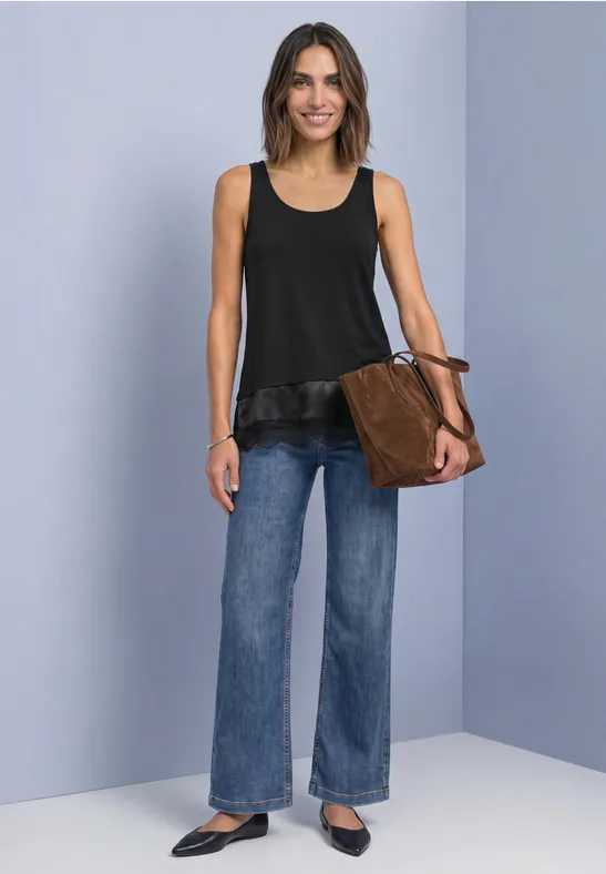Frau in schwarzem ärmellosem Top mit Rüsche, weiten Jeans und brauner Tasche vor grauem Hintergrund.