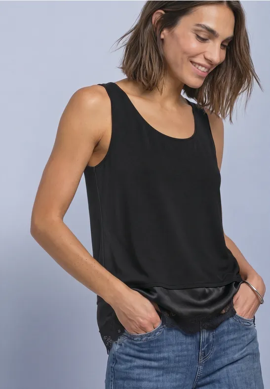 Frau in schwarzem ärmellosem Top mit Satin-Bund und blauen Jeans, die lächelnd die Hände in die Taschen steckt.
