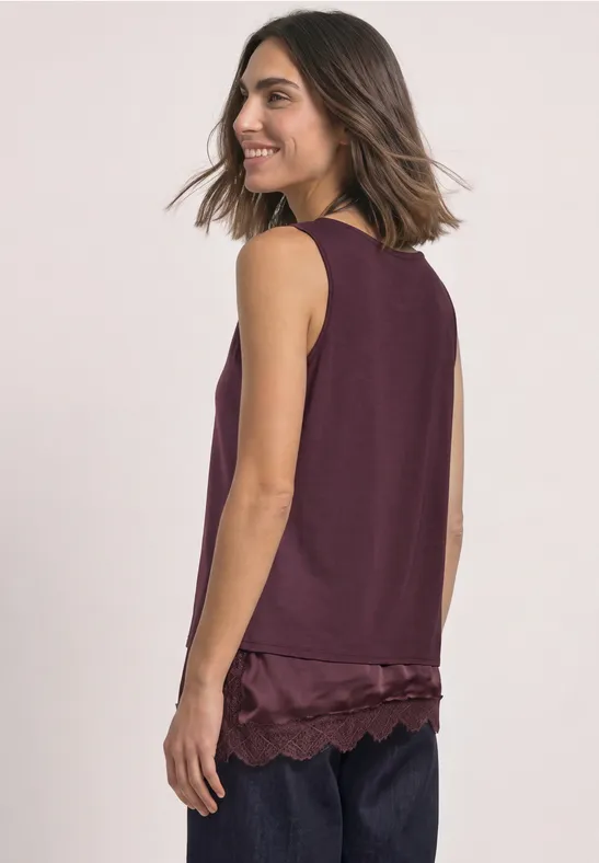 Vrouw in zijaanzicht met een mouwloos bordeauxrood topje met kanten zoom en een donkere broek.