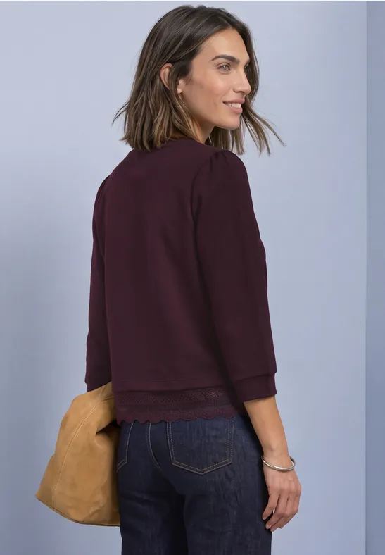 Eine lächelnde Frau in Burgunderjacke und Jeans, seitlich stehend, mit senfgelber Tasche vor grauem Hintergrund.