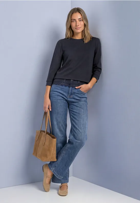 Frau lehnt an grauer Wand, trägt schwarzen Pullover, blaue Jeans, beigen Loafer und braune Tasche.