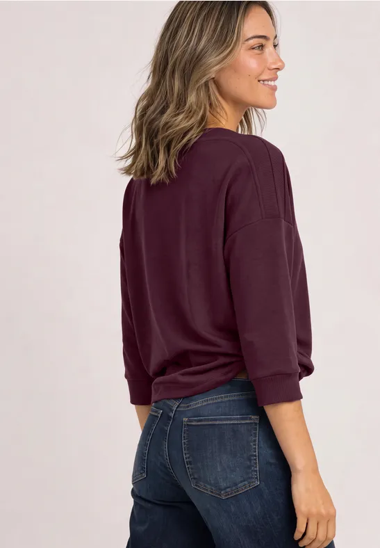 Frau steht seitlich, trägt dunkelroten Pullover und blaue Jeans, lächelt über die Schulter.