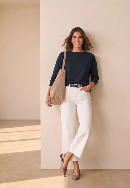 Eine Frau lehnt an einer beigen Wand, trägt schwarzen Pullover, weiße Culottes, beige Tasche und Schuhe, lächelt.